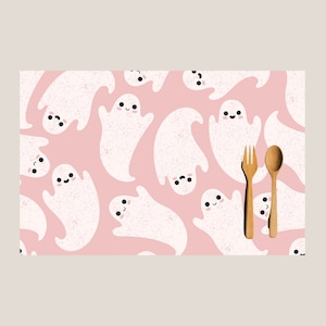 May include: Pink placemat with a white ghost pattern. The ghosts have black eyes and a smiling mouth. There is a wooden fork and spoon on top of the placemat.