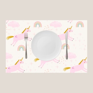 Könnte beinhalten: Ein weißer Platzdeckchen mit einem rosafarbenen Einhorn- und Regenbogen-Design. Das Platzdeckchen hat einen weißen Teller, eine Gabel und ein Messer darauf.