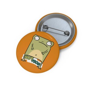 May include: A round pin-back button with an orange background featuring a cartoon illustration of a light green creature with large eyes and the text NOT FROG. The back of the button is silver-colored metal with a horizontal pin mechanism attached.