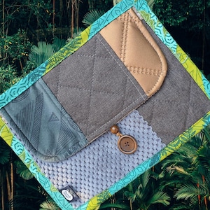 Könnte beinhalten: Eine handgefertigte, gesteppte Clutch mit geometrischem Design. Die Clutch verfügt über eine Vielzahl von Stoffen in Grau-, Blau- und Beigetönen mit einem dekorativen Knopfverschluss. Die Kanten sind mit einem gemusterten Rand in Türkis und Grün verziert.