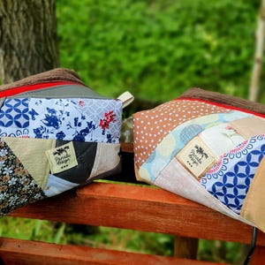 Könnte beinhalten: Zwei rechteckige Patchwork-Reißverschlusstaschen. Eine hat Blumen-, Blau- und Graumuster, die andere Tupfen und blaue geometrische Designs. Beide haben braune Cord-Oberteile, rote Reißverschlüsse und kleine Etiketten.