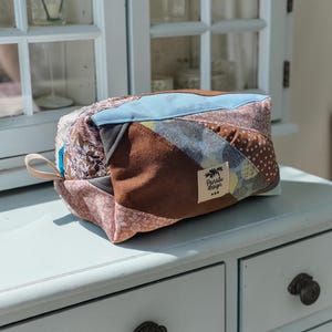 Può includere: Un beauty case patchwork con una varietà di tessuti nei toni del marrone, blu e rosa. La borsa ha una forma rettangolare con chiusura a zip e un piccolo anello. L'etichetta recita "Panabi Design".