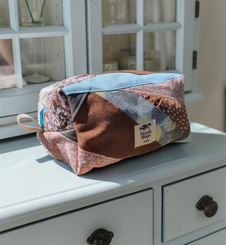 May include: A patchwork cosmetic bag in shades of brown, blue, and pink. The bag has a rectangular shape with a zip closure and a small loop. The bag features a variety of fabrics. The label reads "Panabi Design".