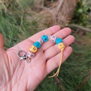 May include: A colourful beaded keychain spelling "SUPERMUM". The beads are yellow, pink, blue and clear. It has a silver clasp and a yellow tassel. The keychain is held in a hand against a blurred green background.