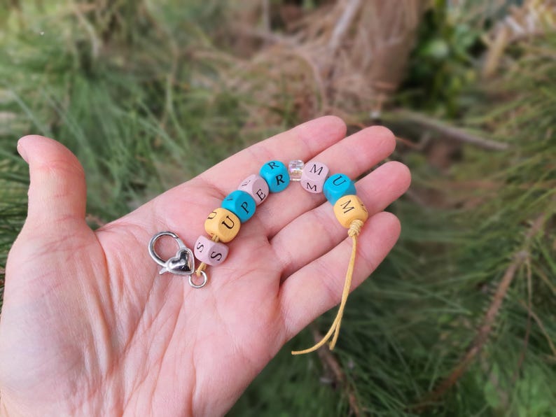 May include: A colourful beaded keychain spelling "SUPERMUM". The beads are yellow, pink, blue and clear. It has a silver clasp and a yellow tassel. The keychain is held in a hand against a blurred green background.