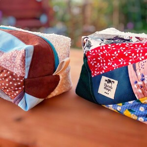 May include: Two cube-shaped bags made from patchwork fabric, featuring a mix of patterns and colours like brown, blue, red, and floral designs. Each bag has a fluffy white top and a small label.