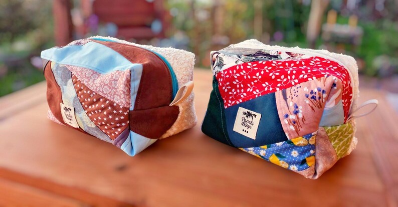 May include: Two cube-shaped bags made from patchwork fabric, featuring a mix of patterns and colours like brown, blue, red, and floral designs. Each bag has a fluffy white top and a small label.
