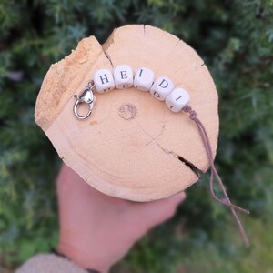May include: A personalised keychain spelling "HEIDI" in white wooden letter beads. The beads are strung on a brown cord and attached to a silver clasp. The keychain is displayed on a light brown wooden slice.