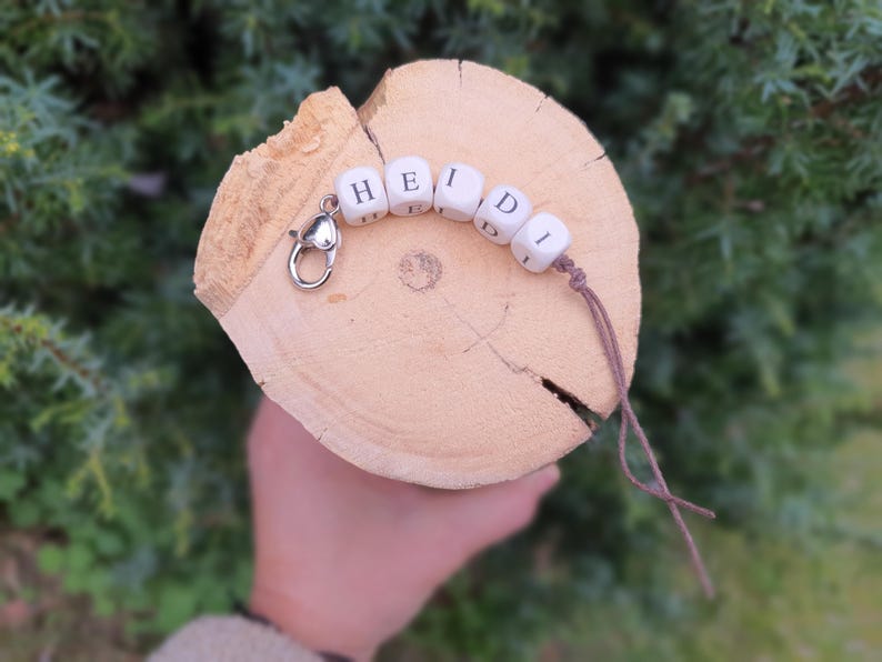 May include: A personalised keychain spelling "HEIDI" in white wooden letter beads. The beads are strung on a brown cord and attached to a silver clasp. The keychain is displayed on a light brown wooden slice.