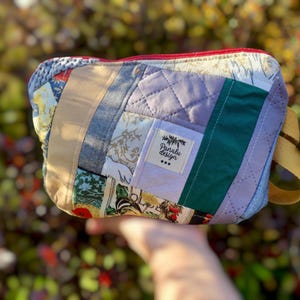 Könnte beinhalten: Ein Patchwork-Etui mit Reißverschluss, rotem Reißverschluss und gelbem Riemen. Das Etui besteht aus verschiedenen Stoffresten in Blau-, Grün-, Beige- und Lilatönen. Ein kleines Etikett trägt die Aufschrift "Panalu design".