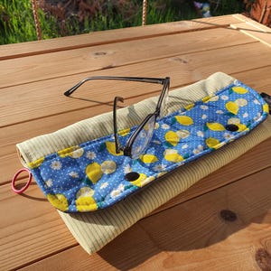 Könnte beinhalten: Eine Brille liegt auf einem gepolsterten Brillenetui. Das Etui hat einen blauen Stoff mit Zitronenmuster und eine hellgrüne Cord-Rückseite. Ein rotes Gummiband ist an der Seite befestigt. Das Etui ist etwa 18 cm lang.