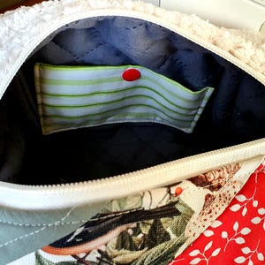 May include: An open bag with a white, fluffy exterior and a zip closure. The interior is lined with dark blue quilted fabric and includes a pocket with green and white stripes, secured by a red button. The exterior features patchwork with floral and red leaf patterns.