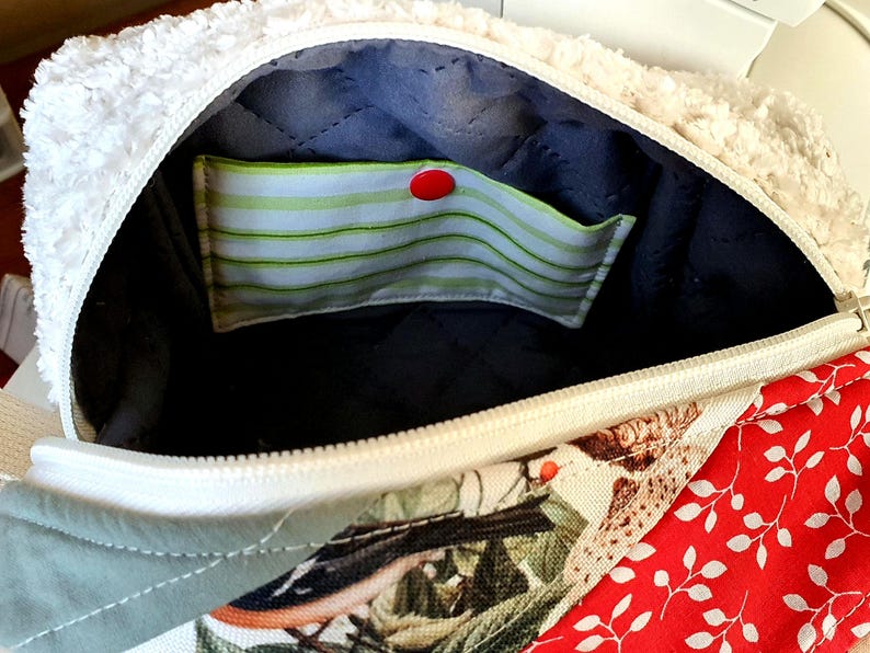 May include: An open bag with a white, fluffy exterior and a zip closure. The interior is lined with dark blue quilted fabric and includes a pocket with green and white stripes, secured by a red button. The exterior features patchwork with floral and red leaf patterns.