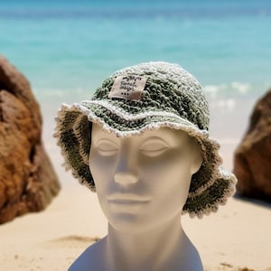 Op de afbeelding: Een gehaakte bucket hat in saliegroen en crèmetinten, voorzien van een golvende, geschulpte rand met een crèmekleurige afwerking. Aan de voorzijde is een klein, rechthoekig stoffen label genaaid met de tekst Rotali Design in zwarte inkt.