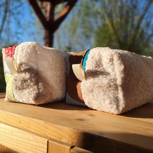 May include: Two rectangular cosmetic bags with a fluffy, textured exterior. One bag has a patchwork design with floral and patterned fabrics, the other has a brown panel. Both have beige ribbon loops. They are set on a wooden surface.