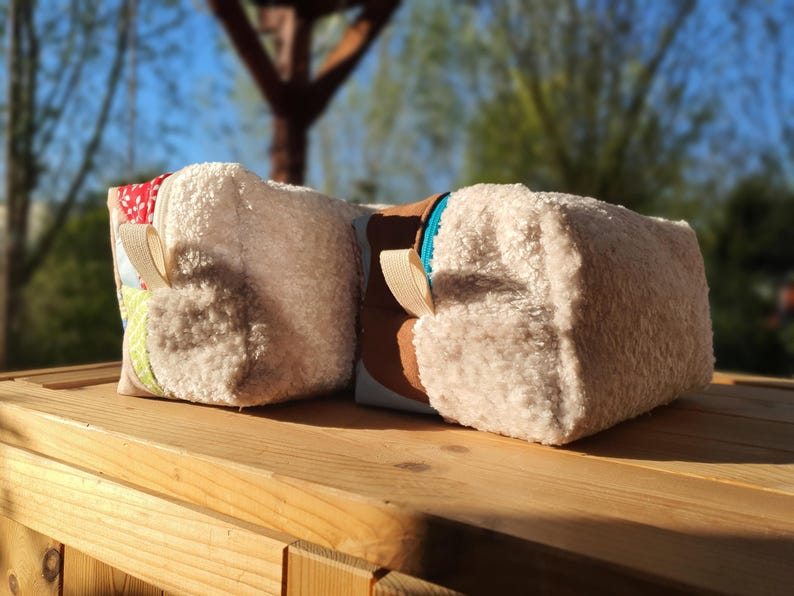 May include: Two rectangular cosmetic bags with a fluffy, textured exterior. One bag has a patchwork design with floral and patterned fabrics, the other has a brown panel. Both have beige ribbon loops. They are set on a wooden surface.