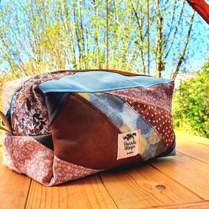 May include: A patchwork travel bag in shades of brown, pink, blue, and tan. The bag features various patterned fabrics, a light blue top, a brown handle, and a label reading "Paralu design".