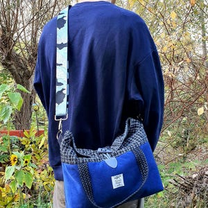 Könnte beinhalten: Blaue Umhängetasche mit gemustertem Riemen und gewebtem Oberteil. Die Tasche hat eine rechteckige Form mit Klappenverschluss und einem kleinen Etikett. Der Riemen hat ein schwarz-weißes Design.