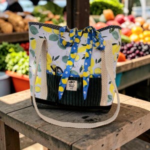 Puede incluir: Un bolso tote acolchado con estampado de limones, con un bolsillo de pana verde oscuro y correas trenzadas color crema. El bolso tiene una cinta azul con detalles de limón y una etiqueta que dice "Panala Design".