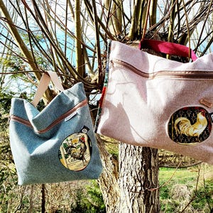 Könnte beinhalten: Zwei Tragetaschen hängen an einem Ast. Eine ist hellblau mit einem Hahn-Emblem, die andere ist beige mit einem Hahn-Emblem. Beide Taschen haben einen Reißverschluss und einen Griff.