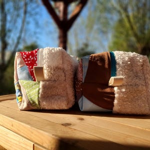 May include: Two small, square fabric pouches with a patchwork design. One features red floral fabric, the other brown. Both have a fluffy, cream-coloured side and a small, tan loop. They sit on a wooden surface.
