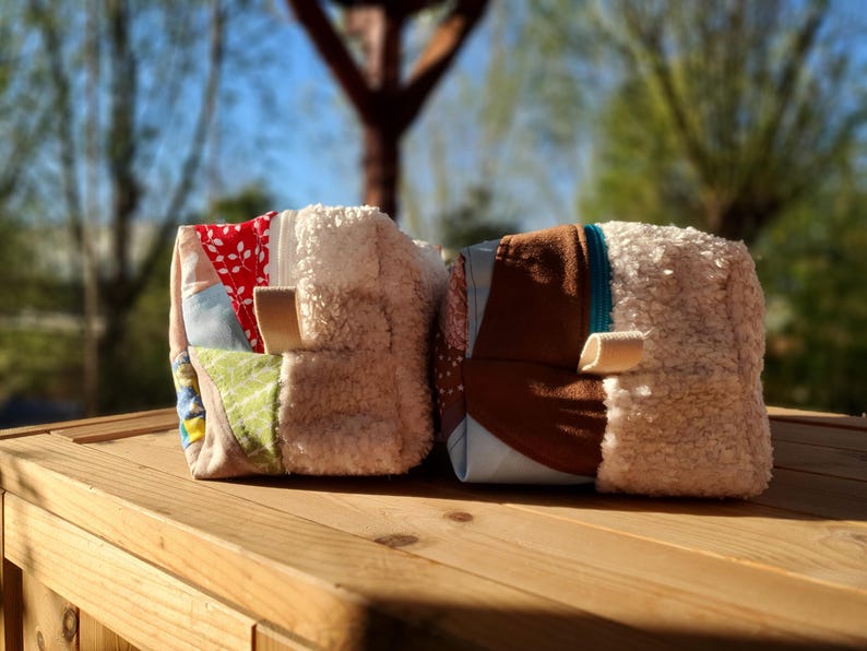 May include: Two small, square fabric pouches with a patchwork design. One features red floral fabric, the other brown. Both have a fluffy, cream-coloured side and a small, tan loop. They sit on a wooden surface.