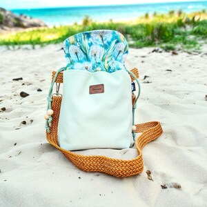 Könnte beinhalten: Hellgrüner Kordelzugbeutel mit gewebtem braunen Boden und Riemen, auf einem Sandstrand. Die Oberseite der Tasche weist ein Blumenmuster mit blauen und weißen Löwenzahnpflanzen auf. Ein kleines braunes Lederetikett ist vorne angebracht.