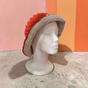 Può includere: Un cappello a secchiello all'uncinetto caratterizzato da un vivace motivo a blocchi di colore con sezioni rosa, arancioni e rosso scuro. Il cappello è rifinito con una tesa larga color beige e una piccola etichetta rettangolare in tessuto.