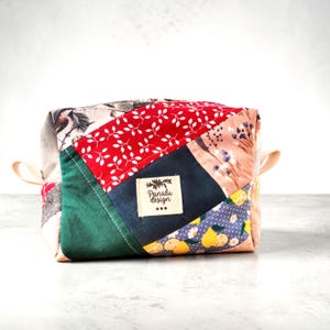 May include: A patchwork fabric box with various patterns and colours. The box features red fabric with white leaves, a green panel, and a navy blue panel with a label reading "Panalu design". Beige handles are attached to the sides.