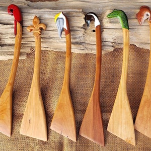 May include: Seven wooden kitchen spatulas with different animal carvings on the handles. The carvings include a cardinal, a fleur-de-lis, a bird, a penguin, a snake, and a fox.