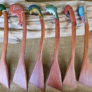 May include: A collection of six wooden spatulas with carved animal and aquatic creature handles. The handles feature a duck, flamingo, fish, shrimp, and a bird. The spatulas are made of a reddish-brown wood.