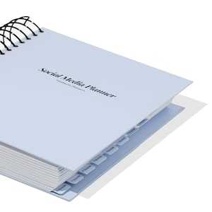 May include: A pale blue spiral-bound planner with the text "Social Media Planner" and "stillness Planners" printed on the cover. The planner has monthly tabs. The spiral binding is black.