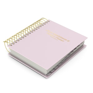 Peut inclure: Un agenda à spirales rose clair avec une reliure en fil doré. La couverture porte le texte doré en relief "Client Appointment & Finance Planner". L'agenda comprend des onglets latéraux pour l'organisation mensuelle et des pages blanches.