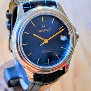 Könnte beinhalten: Eine automatische Bulova Armbanduhr mit dunkelblauem Zifferblatt, goldenen Zeigern und Datumsfenster bei 3 Uhr. Die Uhr hat ein silberfarbenes Gehäuse und ein schwarzes Lederarmband. Das Zifferblatt zeigt "Bulova" und "Automatic 25 Jewels".