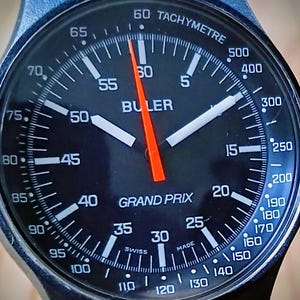 Könnte beinhalten: Nahaufnahme einer Buler Grand Prix Uhr mit schwarzem Zifferblatt, weißen Ziffern und einem orangefarbenen Sekundenzeiger. Die Uhr verfügt über eine Tachymeterskala und ist mit "Swiss Made" gekennzeichnet.