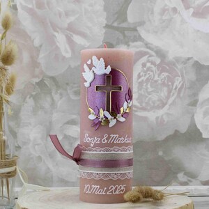 Könnte beinhalten: Eine rosa Kerze mit einem weißen Kreuz und zwei weißen Tauben auf einem violetten Hintergrund. Die Kerze ist mit einem goldenen Kranz und den Namen "Sonya & Markus" sowie dem Datum "10.Mai 2025" verziert.