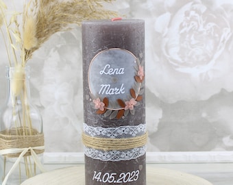 May include: A gray pillar candle with a white lace ribbon and twine. The candle has a floral wreath design with the names "Lena" and "Mark" and the date "14.05.2023" written on it.
