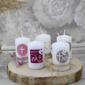 Könnte beinhalten: Fünf weiße Kerzen mit verschiedenen religiösen Symbolen und Farben. Die Kerzen stehen auf einer Holzscheibe. Die Symbole beinhalten ein Kreuz, eine Taube, einen Fisch, einen Anker und ein Herz.
