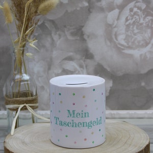 Könnte beinhalten: Weiße Keramik-Sparbüchse mit buntem Punkte-Muster und dem Text "Mein Taschengeld" in Grün. Die Sparbüchse steht auf einer Holzscheibe.