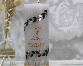 May include: A gray pillar candle with a burlap ribbon and green leaves with gold accents. The candle is personalized with the names "Karla & Julian" and the date "25.02.2019".
