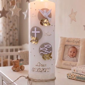 Könnte beinhalten: Eine brennende weiße Säulenkerze, verziert mit religiösen Symbolen, darunter eine Taube, ein Kreuz und Fische. Die Kerze hat goldene Akzente und den Namen "Lirus" mit dem Datum "02.10.2022" aufgedruckt. Die Kerze steht auf einer weißen Oberfläche.