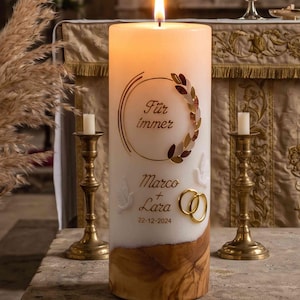 Könnte beinhalten: Eine zylindrische weiße Kerze mit holzfarbenem Fuß, brennender Flamme und der Aufschrift "Für immer", "Marco + Lara" sowie "22-12-2024". Das Design zeigt einen goldenen Lorbeerkranz, zwei weiße Tauben und zwei ineinander verschlungene goldene Ringe.