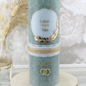 Könnte beinhalten: Eine tealgrüne Hochzeitskerze mit einem weißen Kreis, der die Namen "Luisa" und "Tim" mit drei Herzen enthält. Die Kerze ist mit einem goldenen Blätterkranz und einem braunen und goldenen Bindfadenband verziert. Das Datum "04.05.2023" und zwei goldene Ringe befinden sich ebenfalls auf der Kerze.