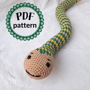 Può includere: Modello di serpente all'uncinetto in PDF. Il serpente è verde e giallo con una testa marrone e un viso sorridente.