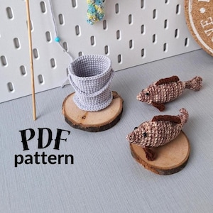 Può includere: Schema a uncinetto per un set a tema pesca, che include un secchio, una canna da pesca e due pesci. Lo schema è disponibile per il download in PDF. I pesci sono marroni e bianchi, e il secchio è grigio.