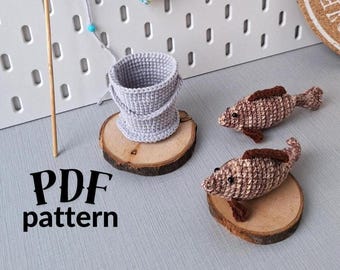 May include: Crochet pattern for a fishing themed set, including a bucket, a fishing rod, and two fish. The pattern is available as a PDF download. The fish are brown and white, and the bucket is gray.