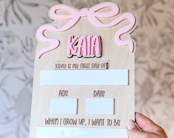 May include: A wooden sign with a pink bow design at the top and the name "KAIA" in pink letters. The sign includes spaces to write "TODAY IS MY FIRST DAY OF", "AGE", "DATE", and "WHEN I GROW UP, I WANT TO BE".