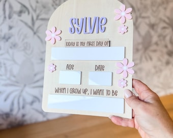 May include: A wooden sign with the name "SYLVIE" in purple letters, adorned with pink flower accents. The sign includes spaces to record the age and date, and the phrase "TODAY IS MY FIRST DAY OF" and "WHEN I GROW UP, I WANT TO BE."