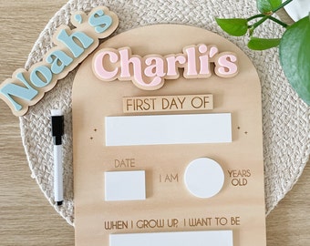 May include: A wooden sign with a light blue and pink nameplate that says "Noah's" and "Charli's". The sign has a white dry-erase surface with the text "LAST DAY OF", "FIRST DAY OF", "DATE", "I AM", "YEARS OLD", and "WHEN I GROW UP, I WANT TO BE".