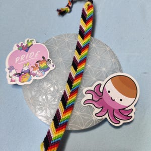 Könnte beinhalten: Ein gewebtes Armband mit Chevron-Muster in Regenbogenfarben wie Rot, Orange, Gelb, Grün, Blau und Violett sowie schwarzen und weißen Fäden. Zwei Aufkleber sind vorhanden: einer mit Hasen und dem Text PRIDE sowie einer mit einem Oktopus.